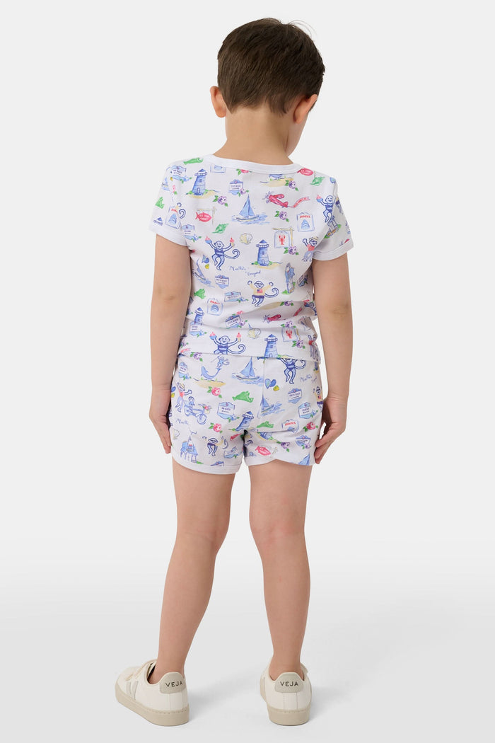 A young child stands barefoot, smiling and facing forward, wearing Roller Rabbit Kids Monkey On The Vineyard Pajamas made of soft Pima cotton with colorful cartoon patterns of animals, vehicles, and buildings. The pajamas feature buttons at the neckline.