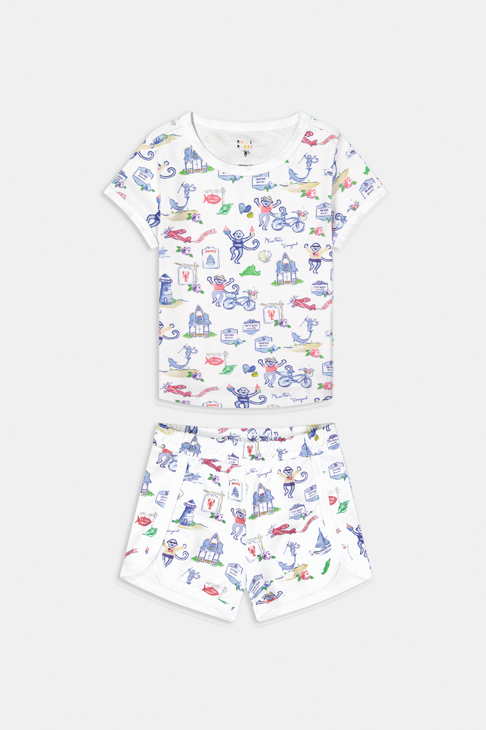 A young child stands barefoot, smiling and facing forward, wearing Roller Rabbit Kids Monkey On The Vineyard Pajamas made of soft Pima cotton with colorful cartoon patterns of animals, vehicles, and buildings. The pajamas feature buttons at the neckline.
