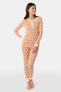 A woman stands barefoot, smiling and looking off to the side, wearing Roller Rabbit Kids Monkey Pajamas—white and orange long-sleeve henley set with playful monkey print and white cuffs.
