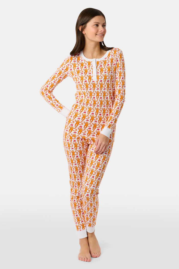A woman stands barefoot, smiling and looking off to the side, wearing Roller Rabbit Kids Monkey Pajamas—white and orange long-sleeve henley set with playful monkey print and white cuffs.
