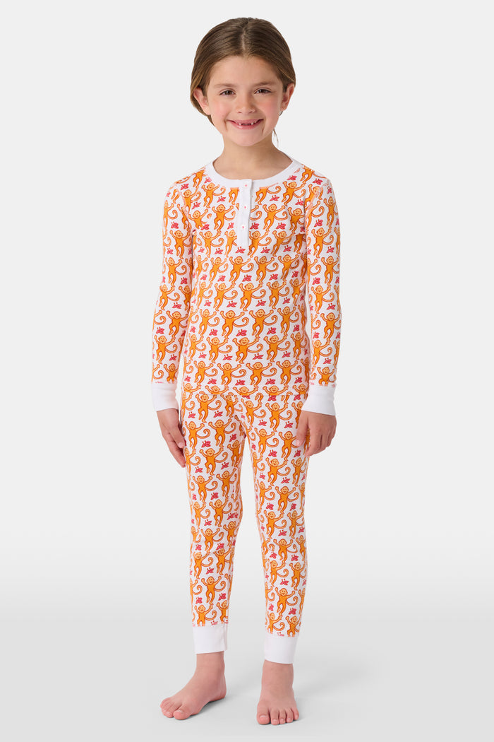 A woman stands barefoot, smiling and looking off to the side, wearing Roller Rabbit Kids Monkey Pajamas—white and orange long-sleeve henley set with playful monkey print and white cuffs.