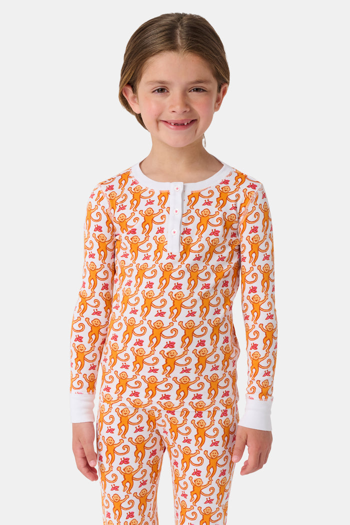 A woman stands barefoot, smiling and looking off to the side, wearing Roller Rabbit Kids Monkey Pajamas—white and orange long-sleeve henley set with playful monkey print and white cuffs.