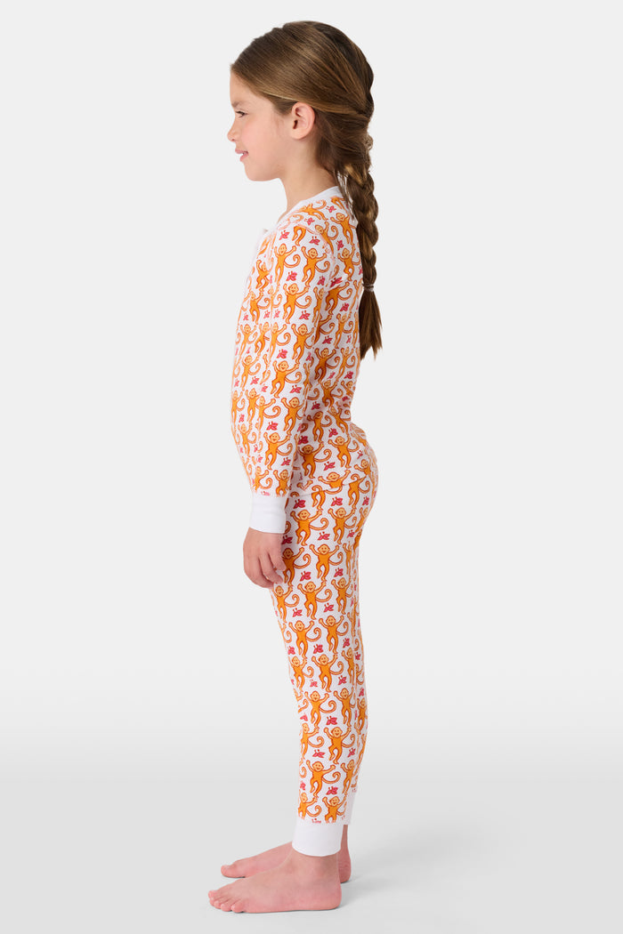 A woman stands barefoot, smiling and looking off to the side, wearing Roller Rabbit Kids Monkey Pajamas—white and orange long-sleeve henley set with playful monkey print and white cuffs.