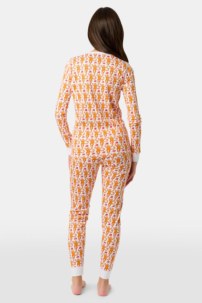 A woman stands barefoot, smiling and looking off to the side, wearing Roller Rabbit Kids Monkey Pajamas—white and orange long-sleeve henley set with playful monkey print and white cuffs.