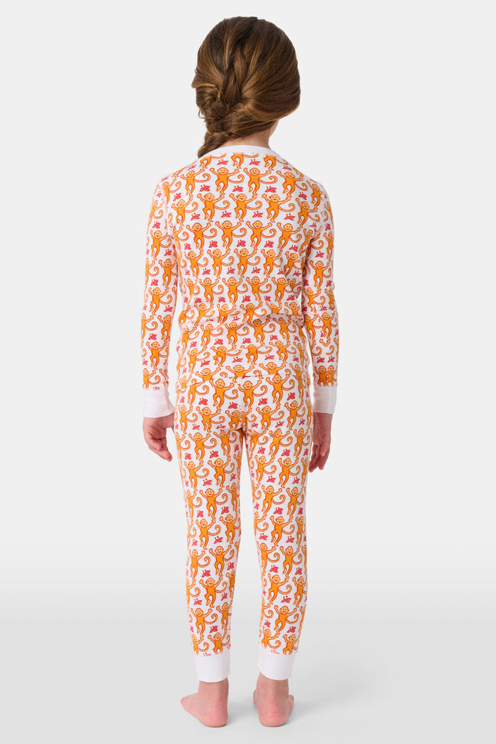 A woman stands barefoot, smiling and looking off to the side, wearing Roller Rabbit Kids Monkey Pajamas—white and orange long-sleeve henley set with playful monkey print and white cuffs.