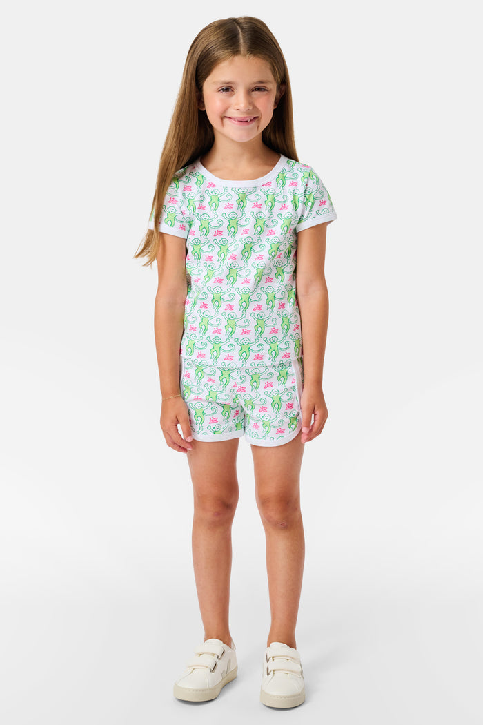 Smiling young girl with long brown hair wearing the Roller Rabbit Kids Monkey Playa Play Set and white shoes, standing against a plain white background.