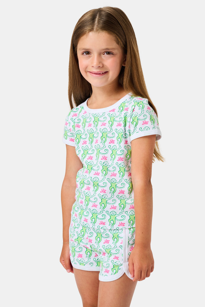 Smiling young girl with long brown hair wearing the Roller Rabbit Kids Monkey Playa Play Set and white shoes, standing against a plain white background.