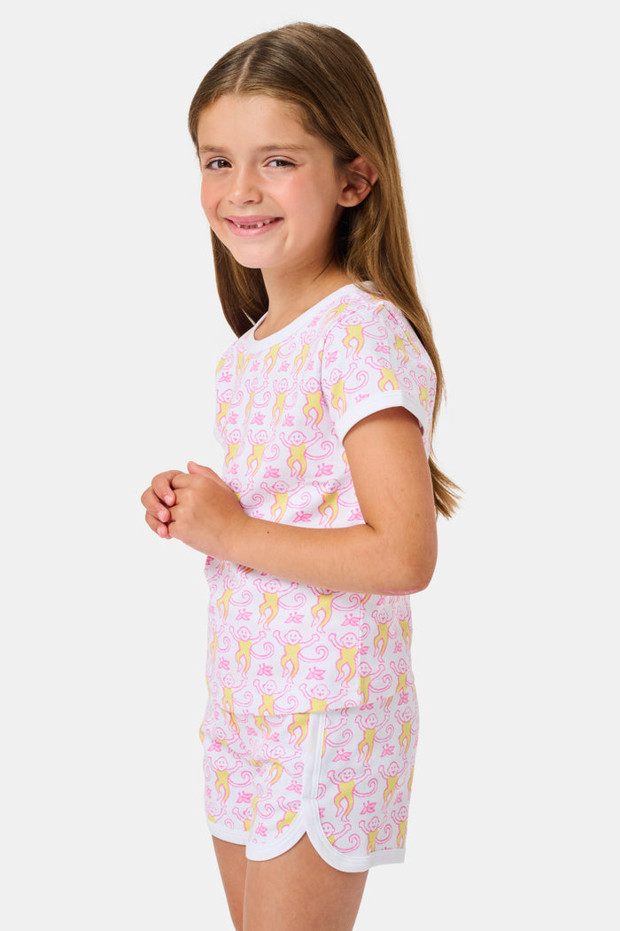 A young girl with long brown hair wears the Roller Rabbit Kids Monkey Playa Play Set and white shoes, standing and smiling against a plain white background.