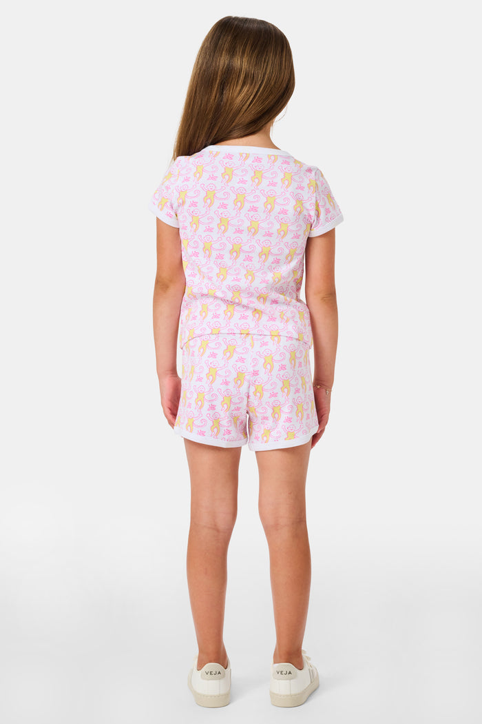A young girl with long brown hair wears the Roller Rabbit Kids Monkey Playa Play Set and white shoes, standing and smiling against a plain white background.