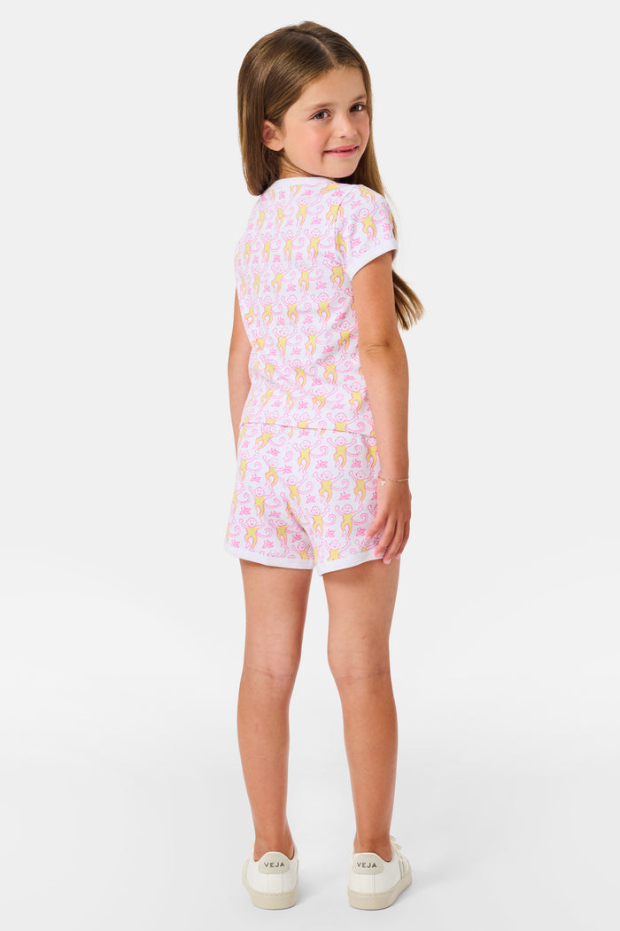 A young girl with long brown hair wears the Roller Rabbit Kids Monkey Playa Play Set and white shoes, standing and smiling against a plain white background.