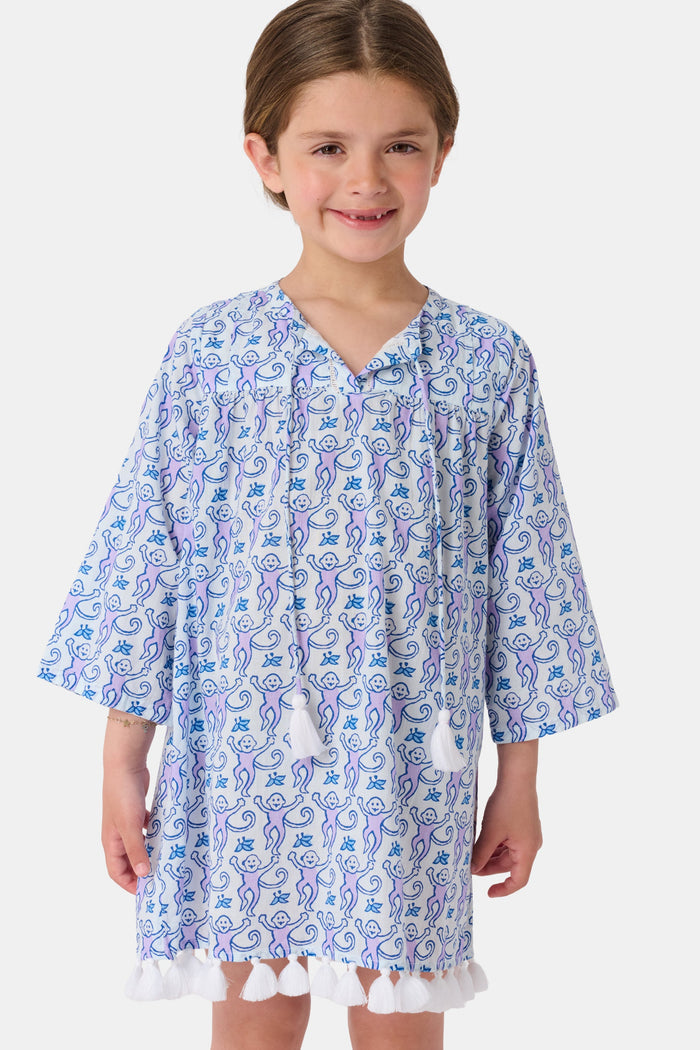 A young girl smiles while wearing the Roller Rabbit Kids Monkey Serafina Tunic, a blue and white patterned cotton voile design with tassels at the hem. She stands facing forward in flip-flops against a plain white background.