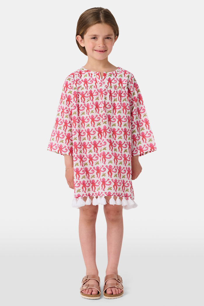 Young girl in a loose Kids Monkey Serafina Tunic by Roller Rabbit—a long-sleeved cotton voile dress with a playful monkey print and white ruffled hem—smiling against a plain white background.