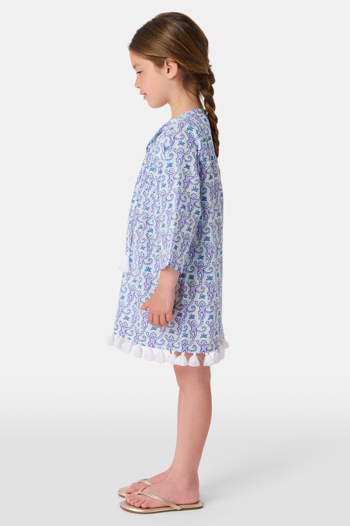 A young girl smiles while wearing the Roller Rabbit Kids Monkey Serafina Tunic, a blue and white patterned cotton voile design with tassels at the hem. She stands facing forward in flip-flops against a plain white background.
