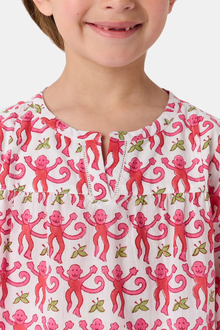 Young girl in a loose Kids Monkey Serafina Tunic by Roller Rabbit—a long-sleeved cotton voile dress with a playful monkey print and white ruffled hem—smiling against a plain white background.