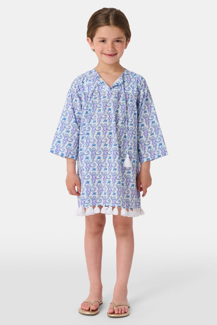 A young girl smiles while wearing the Roller Rabbit Kids Monkey Serafina Tunic, a blue and white patterned cotton voile design with tassels at the hem. She stands facing forward in flip-flops against a plain white background.