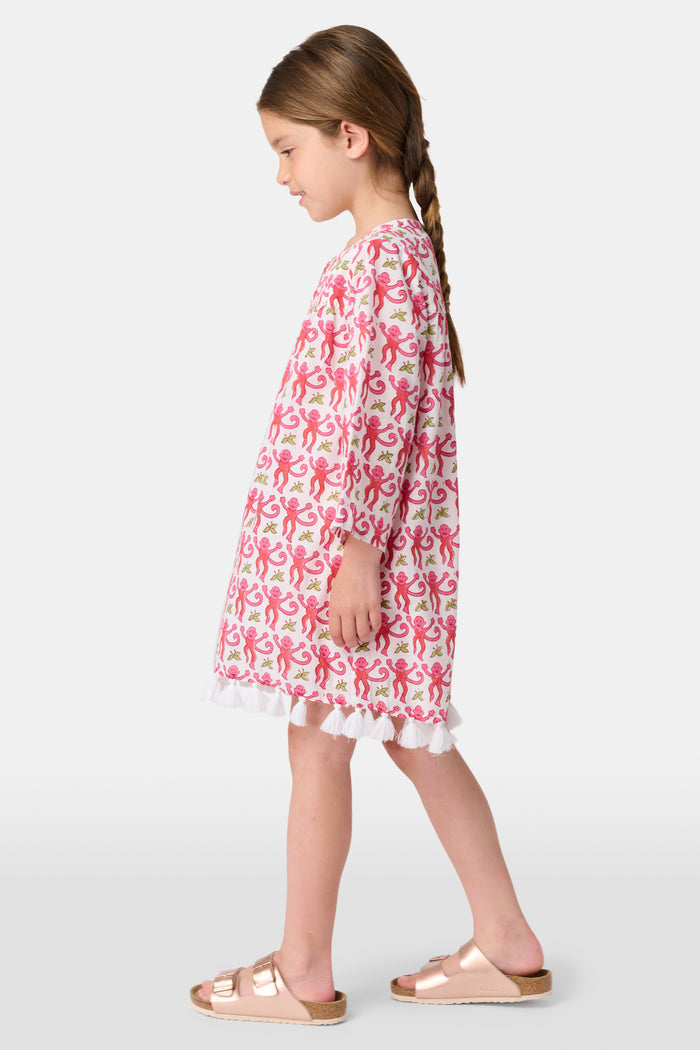 Young girl in a loose Kids Monkey Serafina Tunic by Roller Rabbit—a long-sleeved cotton voile dress with a playful monkey print and white ruffled hem—smiling against a plain white background.