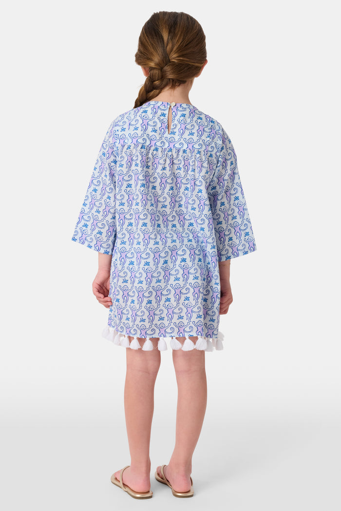 A young girl smiles while wearing the Roller Rabbit Kids Monkey Serafina Tunic, a blue and white patterned cotton voile design with tassels at the hem. She stands facing forward in flip-flops against a plain white background.