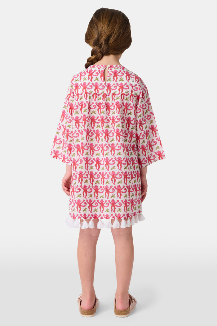 Young girl in a loose Kids Monkey Serafina Tunic by Roller Rabbit—a long-sleeved cotton voile dress with a playful monkey print and white ruffled hem—smiling against a plain white background.