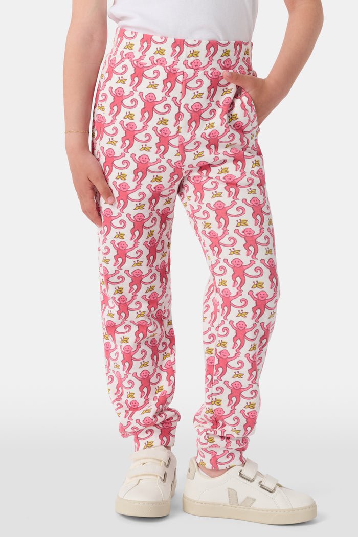 Wearing Roller Rabbit’s Kids Monkey Sweatpants featuring a playful monkey pattern and banded waistband, white sneakers, and a white top, a person stands with one hand in their pocket against a plain background.