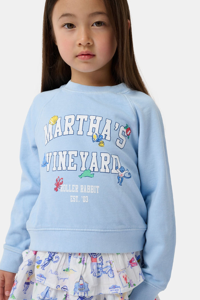 A young girl wears the Kids MV Icons Crewneck by Roller Rabbit—a blue fleece top adorned with playful illustrations—paired with a white skirt featuring colorful prints, standing against a plain background.