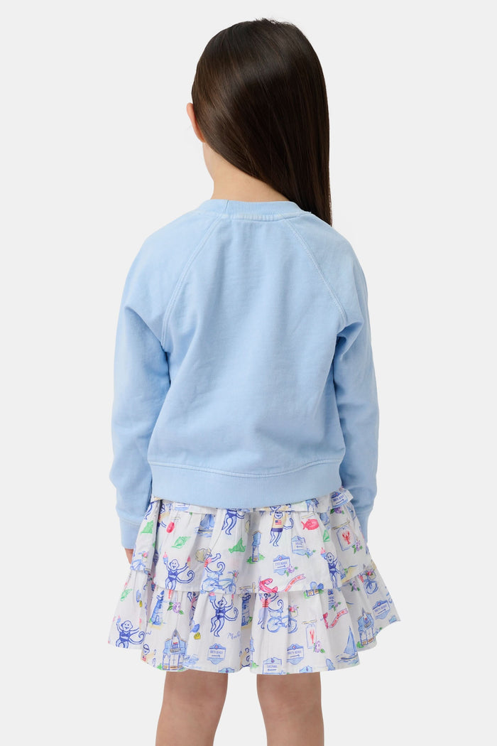 A young girl wears the Kids MV Icons Crewneck by Roller Rabbit—a blue fleece top adorned with playful illustrations—paired with a white skirt featuring colorful prints, standing against a plain background.