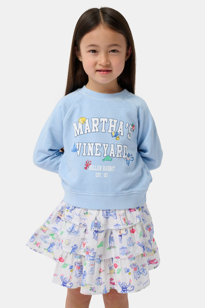 A young girl wears the Kids MV Icons Crewneck by Roller Rabbit—a blue fleece top adorned with playful illustrations—paired with a white skirt featuring colorful prints, standing against a plain background.
