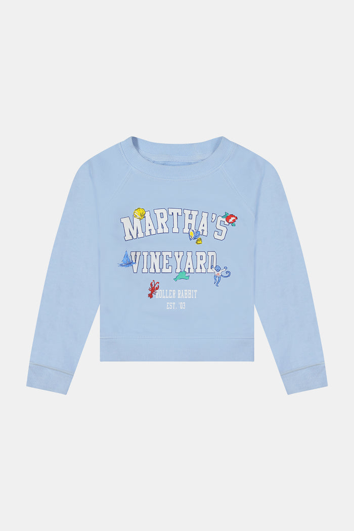 A young girl wears the Kids MV Icons Crewneck by Roller Rabbit—a blue fleece top adorned with playful illustrations—paired with a white skirt featuring colorful prints, standing against a plain background.