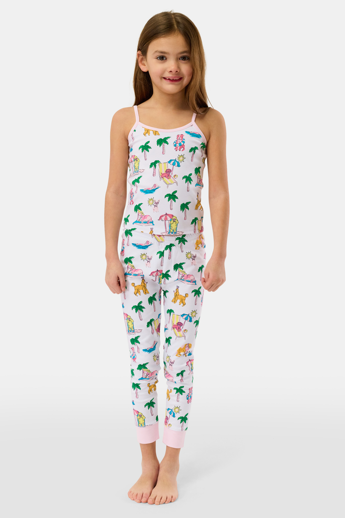 A young girl smiles in the Kids Poolside Pups Noa Set by Roller Rabbit—white Pima Cotton pajamas with colorful animal, palm tree, and pup prints, pink ankle cuffs, and sleeveless thin straps.