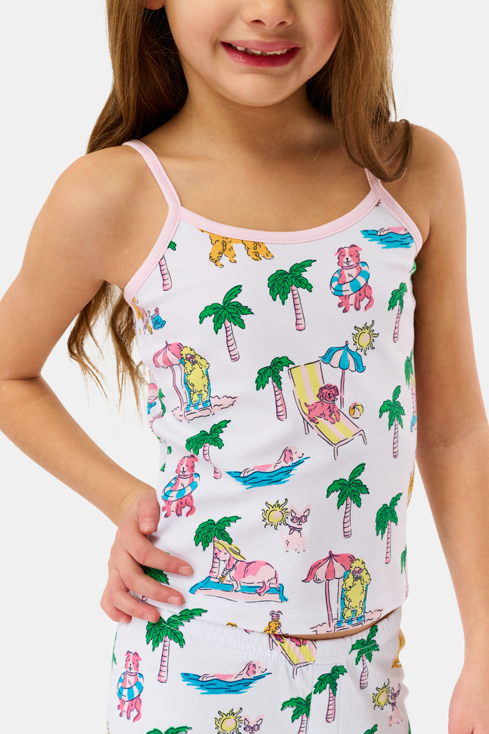 A young girl smiles in the Kids Poolside Pups Noa Set by Roller Rabbit—white Pima Cotton pajamas with colorful animal, palm tree, and pup prints, pink ankle cuffs, and sleeveless thin straps.