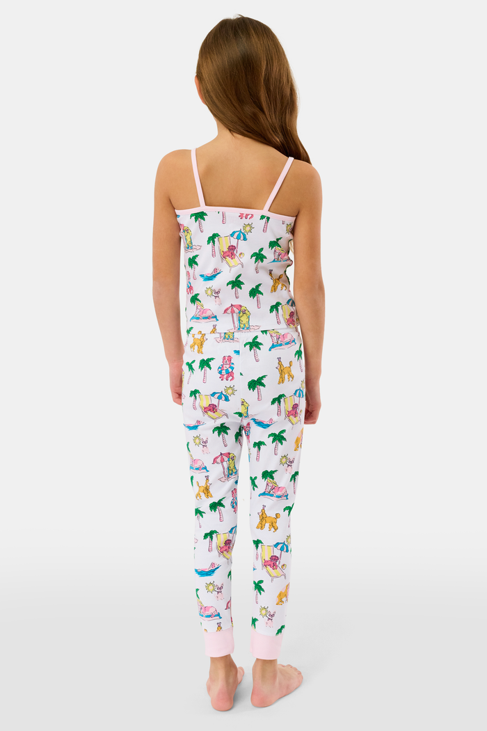 A young girl smiles in the Kids Poolside Pups Noa Set by Roller Rabbit—white Pima Cotton pajamas with colorful animal, palm tree, and pup prints, pink ankle cuffs, and sleeveless thin straps.
