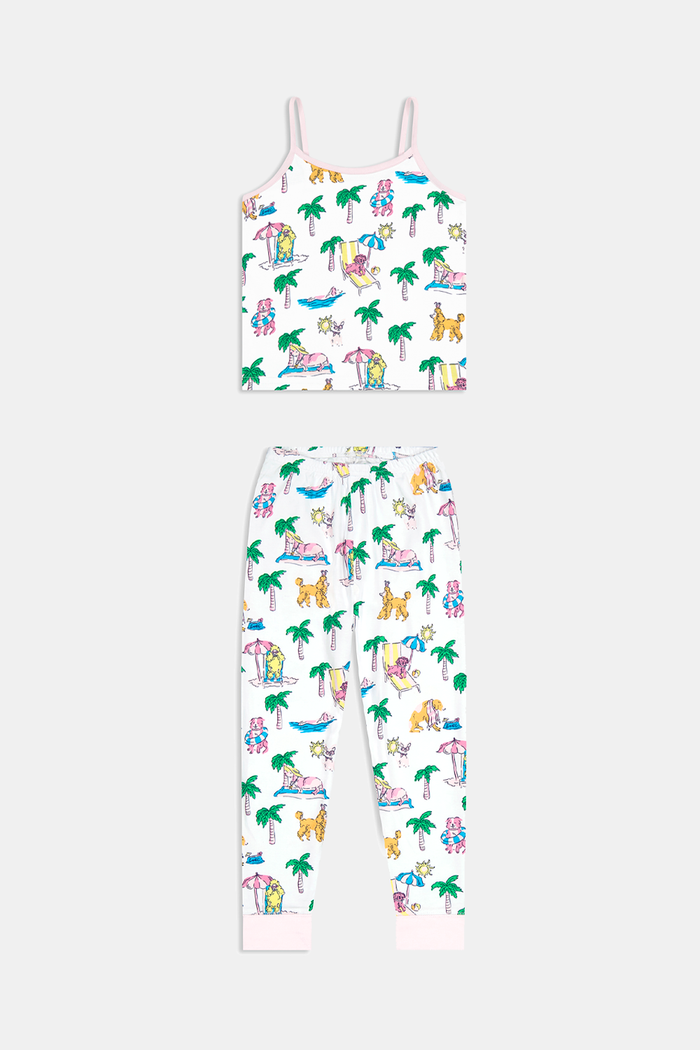 A young girl smiles in the Kids Poolside Pups Noa Set by Roller Rabbit—white Pima Cotton pajamas with colorful animal, palm tree, and pup prints, pink ankle cuffs, and sleeveless thin straps.