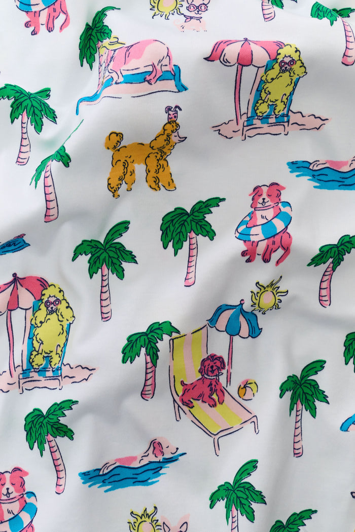 A woman stands barefoot in the Roller Rabbit Poolside Pups Joanie Pajamas, a white Pima Cotton set featuring colorful tropical prints, a sleeveless top with thin straps, and loose wide-leg pants.