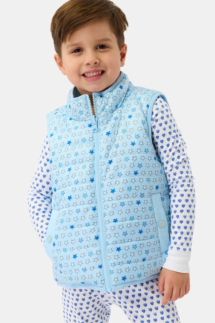 A young child smiles in a Roller Rabbit Kids Pop Stars Kiana Reversible Puffer Vest in sky blue over white pajamas with blue hearts, the vibrant vest contrasting with their short brown hair and the plain light background.