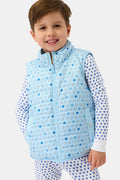 A young child smiles in a Roller Rabbit Kids Pop Stars Kiana Reversible Puffer Vest in sky blue over white pajamas with blue hearts, the vibrant vest contrasting with their short brown hair and the plain light background.