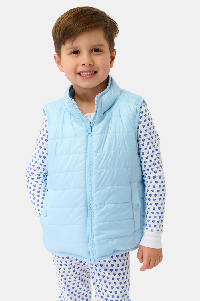 A young child smiles in a Roller Rabbit Kids Pop Stars Kiana Reversible Puffer Vest in sky blue over white pajamas with blue hearts, the vibrant vest contrasting with their short brown hair and the plain light background.