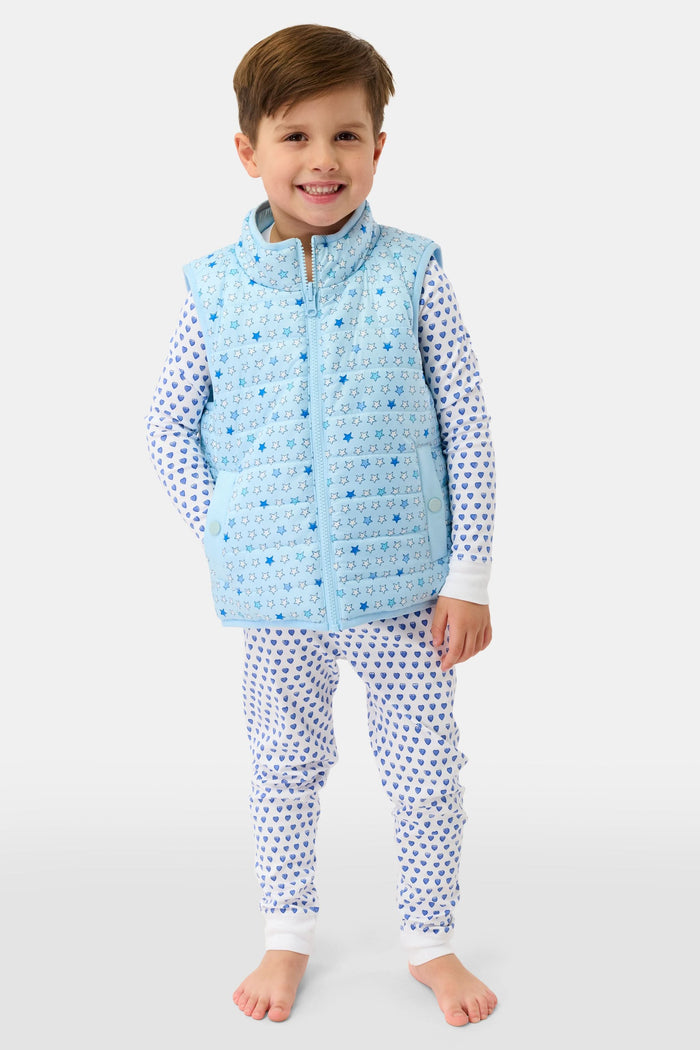 A young child smiles in a Roller Rabbit Kids Pop Stars Kiana Reversible Puffer Vest in sky blue over white pajamas with blue hearts, the vibrant vest contrasting with their short brown hair and the plain light background.