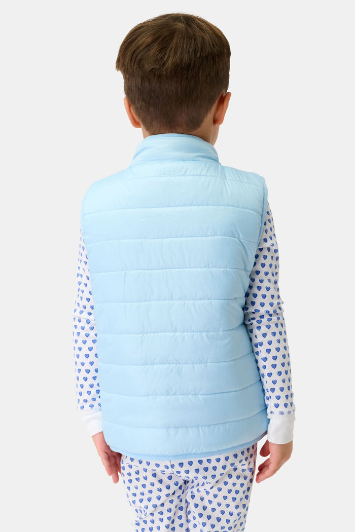 A young child smiles in a Roller Rabbit Kids Pop Stars Kiana Reversible Puffer Vest in sky blue over white pajamas with blue hearts, the vibrant vest contrasting with their short brown hair and the plain light background.