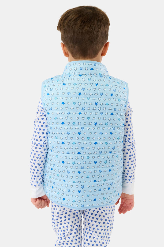 A young child smiles in a Roller Rabbit Kids Pop Stars Kiana Reversible Puffer Vest in sky blue over white pajamas with blue hearts, the vibrant vest contrasting with their short brown hair and the plain light background.