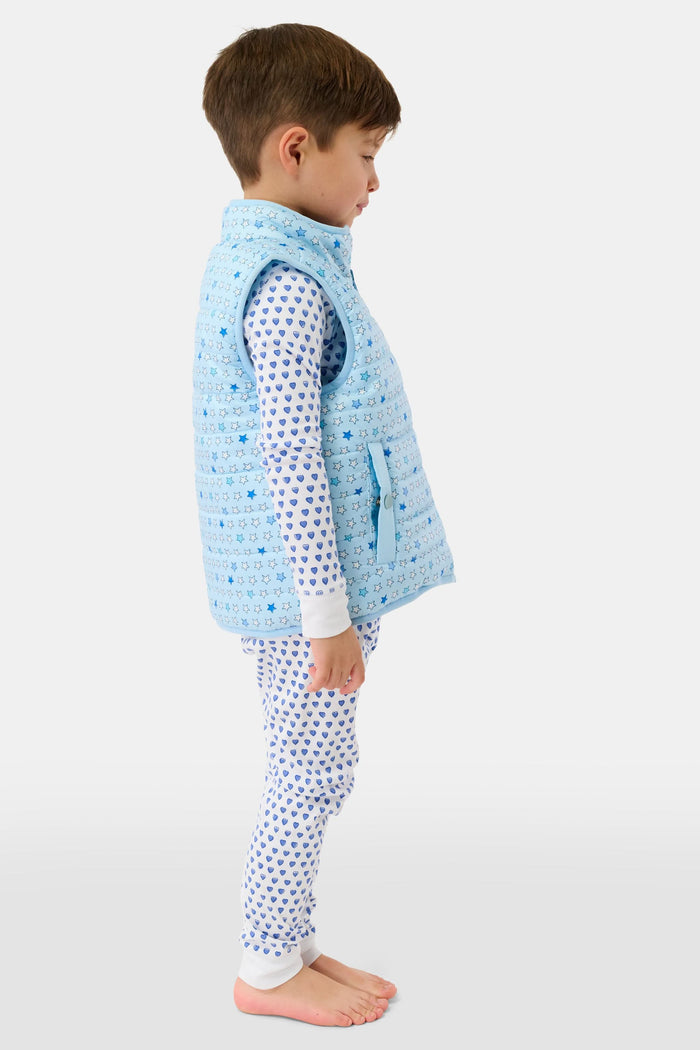 A young child smiles in a Roller Rabbit Kids Pop Stars Kiana Reversible Puffer Vest in sky blue over white pajamas with blue hearts, the vibrant vest contrasting with their short brown hair and the plain light background.