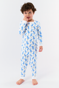 A young child with curly hair stands barefoot in white Kids Romy Rabbit Pajamas by Roller Rabbit, featuring a blue bunny print, looking at the camera and touching their mouth with one finger.