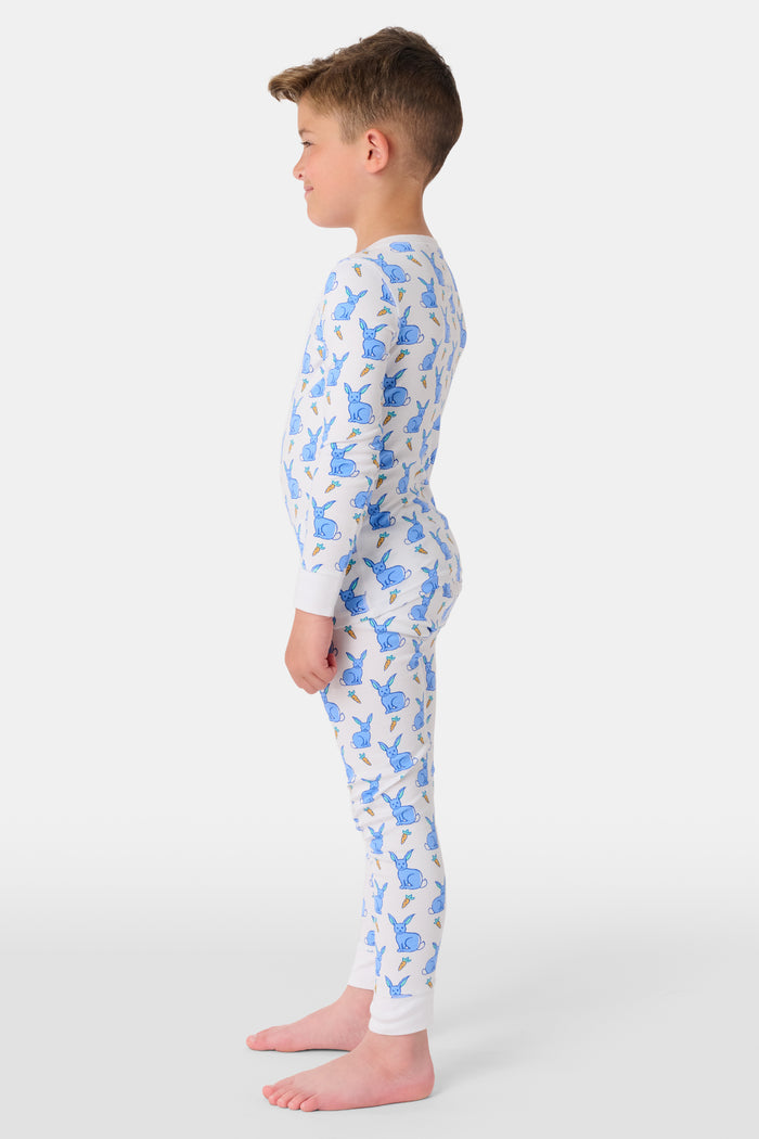 A young child with curly hair stands barefoot in white Kids Romy Rabbit Pajamas by Roller Rabbit, featuring a blue bunny print, looking at the camera and touching their mouth with one finger.