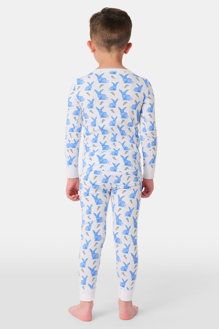 A young child with curly hair stands barefoot in white Kids Romy Rabbit Pajamas by Roller Rabbit, featuring a blue bunny print, looking at the camera and touching their mouth with one finger.