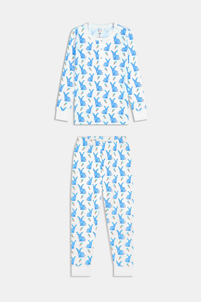 A young child with curly hair stands barefoot in white Kids Romy Rabbit Pajamas by Roller Rabbit, featuring a blue bunny print, looking at the camera and touching their mouth with one finger.