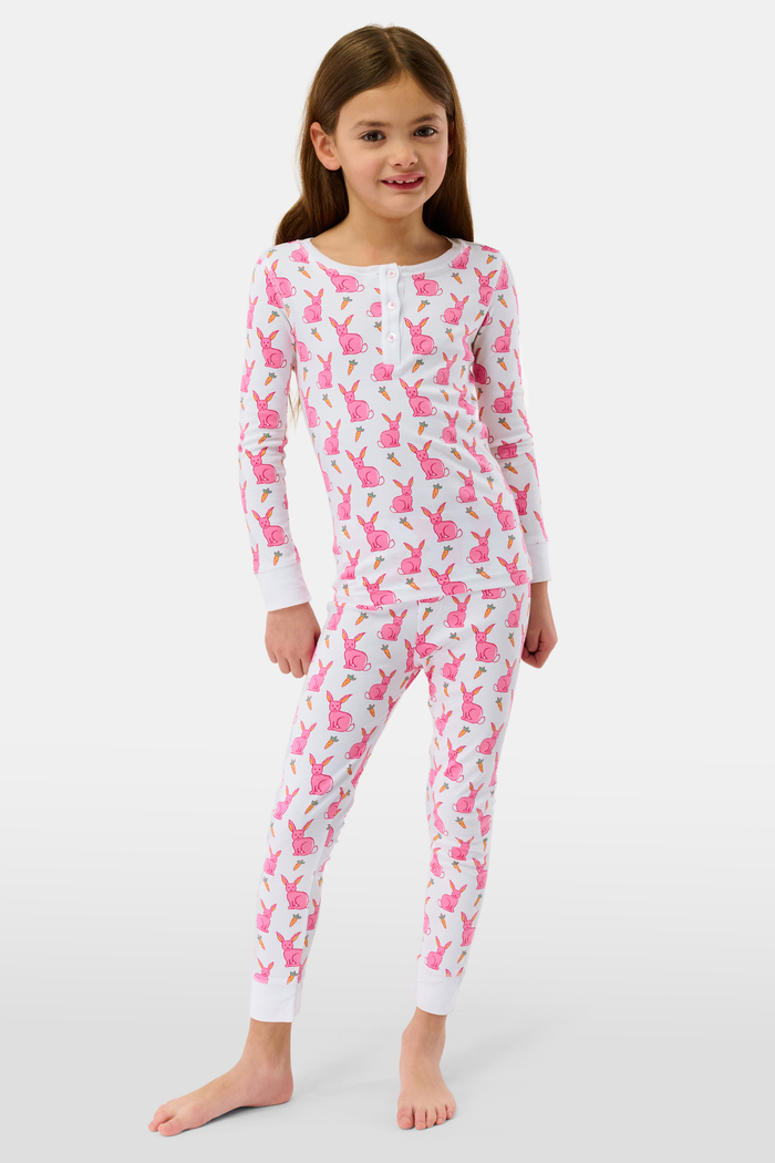 A young girl smiles barefoot in Roller Rabbit’s Kids Romy the Rabbit Pajamas, a soft Pima cotton long-sleeve set with a playful rabbit print; her brown hair frames her face against a simple, light background.