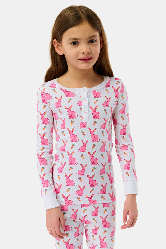 A young girl smiles barefoot in Roller Rabbit’s Kids Romy the Rabbit Pajamas, a soft Pima cotton long-sleeve set with a playful rabbit print; her brown hair frames her face against a simple, light background.