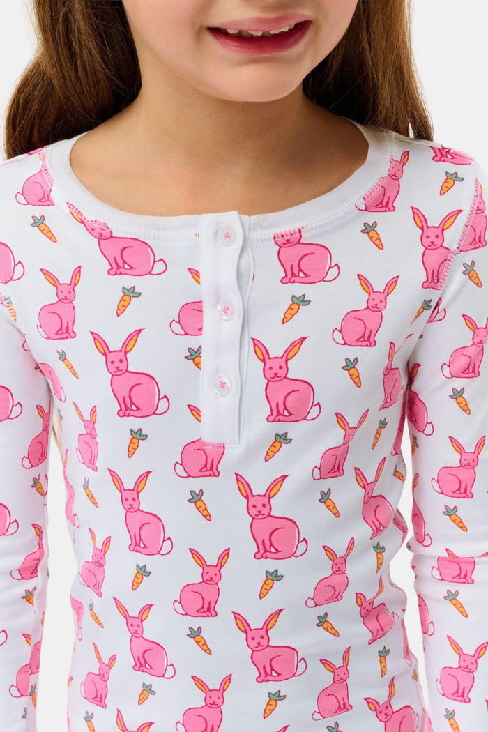 A young girl smiles barefoot in Roller Rabbit’s Kids Romy the Rabbit Pajamas, a soft Pima cotton long-sleeve set with a playful rabbit print; her brown hair frames her face against a simple, light background.