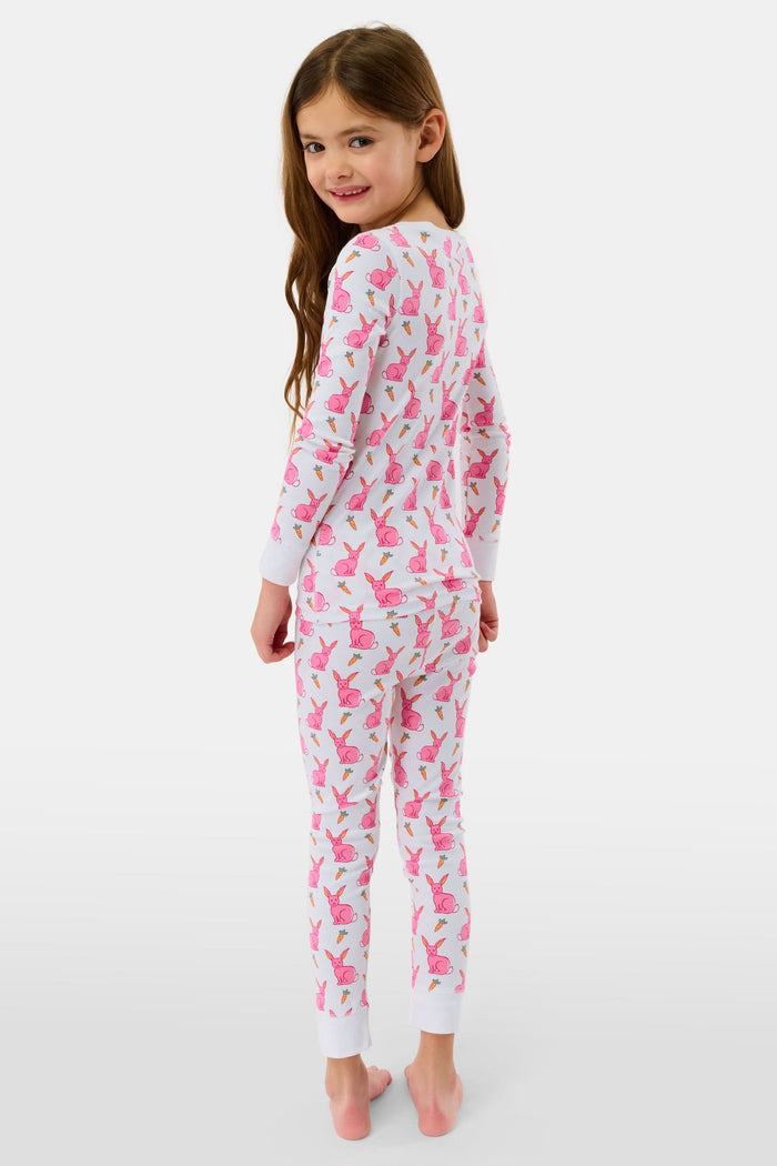 A young girl smiles barefoot in Roller Rabbit’s Kids Romy the Rabbit Pajamas, a soft Pima cotton long-sleeve set with a playful rabbit print; her brown hair frames her face against a simple, light background.
