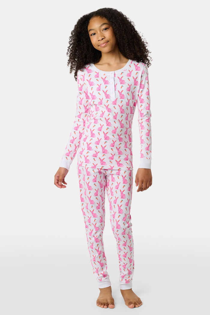 A young girl smiles barefoot in Roller Rabbit’s Kids Romy the Rabbit Pajamas, a soft Pima cotton long-sleeve set with a playful rabbit print; her brown hair frames her face against a simple, light background.