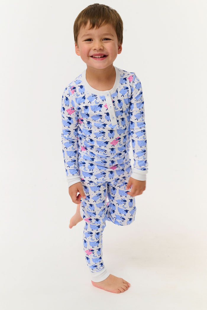 A young boy smiles on a white backdrop, wearing Roller Rabbit's Kids Sammy The Sheep Pajamas—cozy Pima cotton sleepwear featuring a playful sheep print, perfect for sweet dreams.