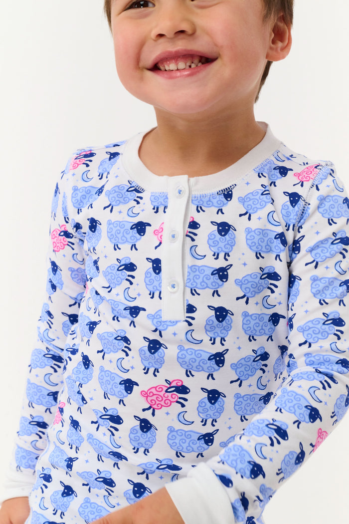 A young boy smiles on a white backdrop, wearing Roller Rabbit's Kids Sammy The Sheep Pajamas—cozy Pima cotton sleepwear featuring a playful sheep print, perfect for sweet dreams.